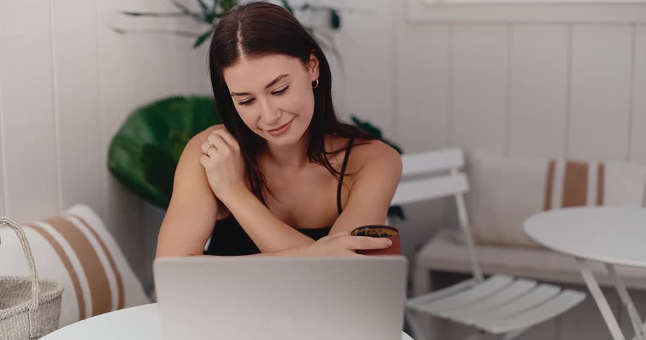 A young woman smiles while looking at her laptop screen and holding a coffee mug in a bright, modern cafe