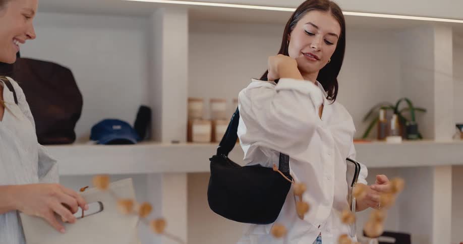 A stylish young woman excitedly tries on a black shoulder bag with her friend in a contemporary fashion boutique