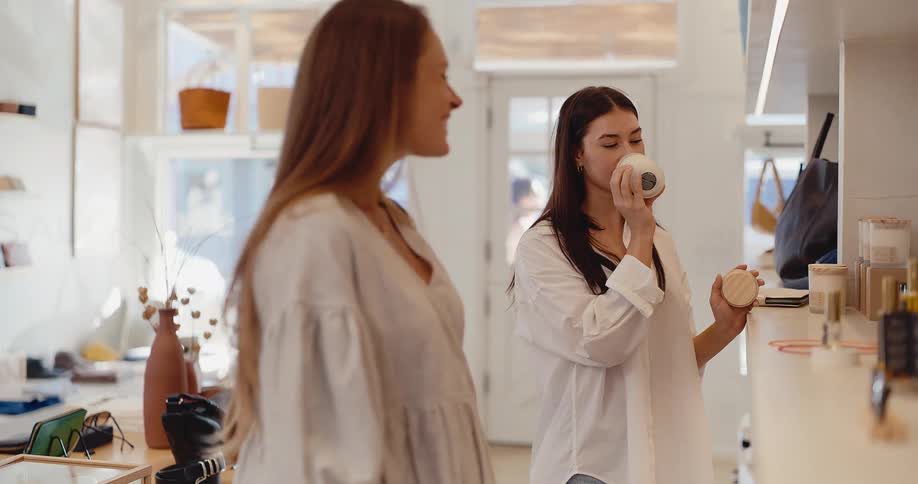 Two stylish young women enjoy smelling scented candles while shopping together in a chic modern boutique