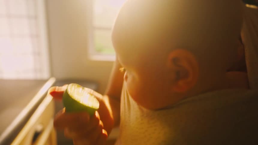 A curious baby in a sling inspects a fresh lime held by its mother in a sunlit kitchen