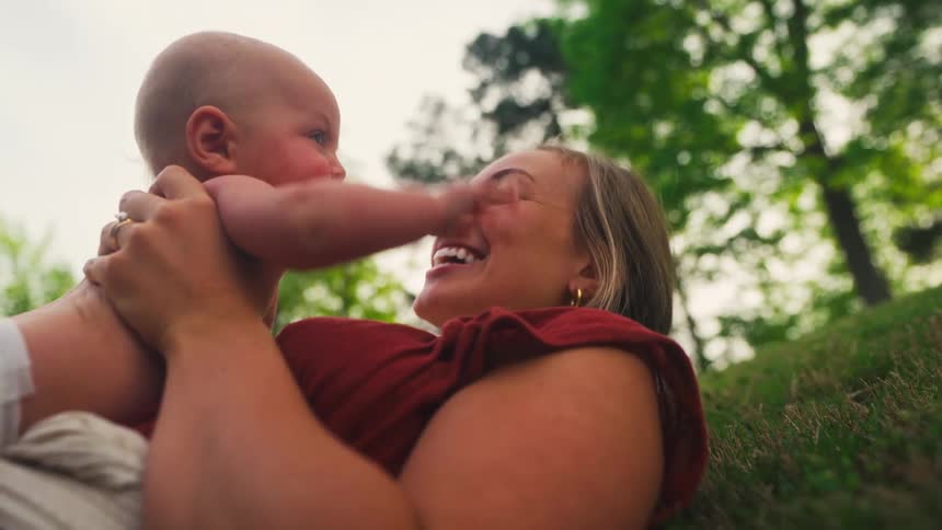 A joyful mother lies on the grass in a park, laughing and playing with her adorable newborn baby