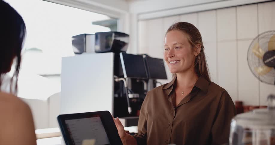 A smiling barista turns the POS terminal for a customer to pay with a credit card in a bright, modern cafe