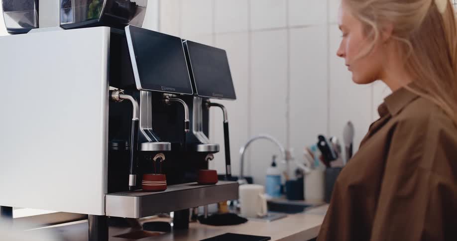 A professional female barista carefully watches as a modern espresso machine brews fresh coffee into two cups in a bright and clean cafe
