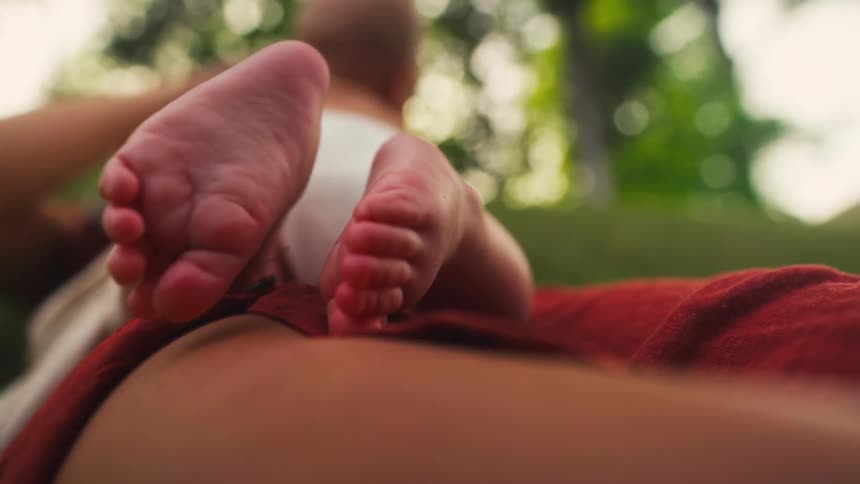 A close-up shot focuses on the tiny feet of a newborn baby resting on its mother's lap while they relax together in a park