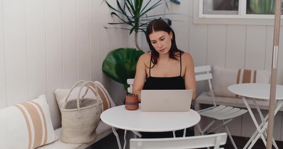 Young female freelancer works on her laptop at a minimalist white cafe table outdoors