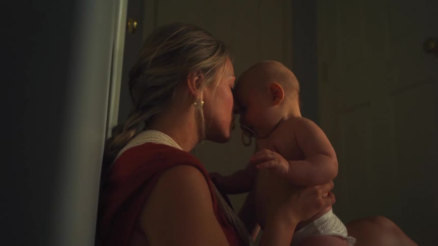 A mother tenderly holds her baby, smiling and kissing their forehead in a softly lit room