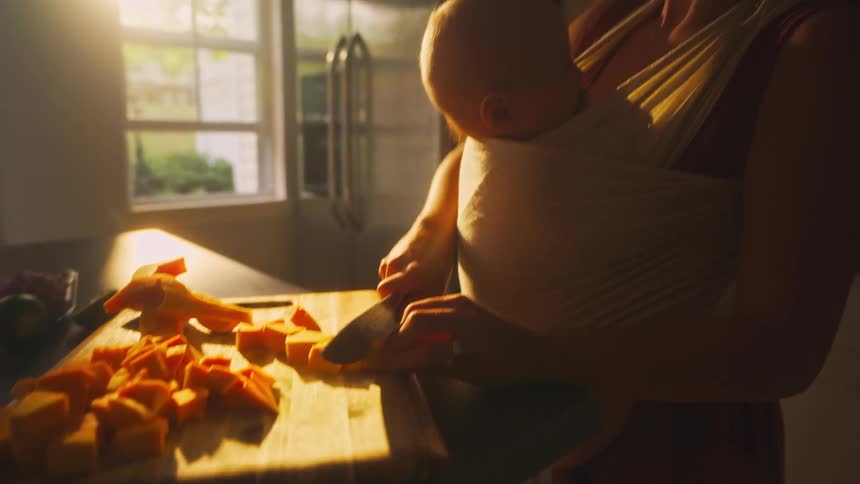 A mother carries her baby in a sling while chopping vegetables on a cutting board in a sun-drenched kitchen