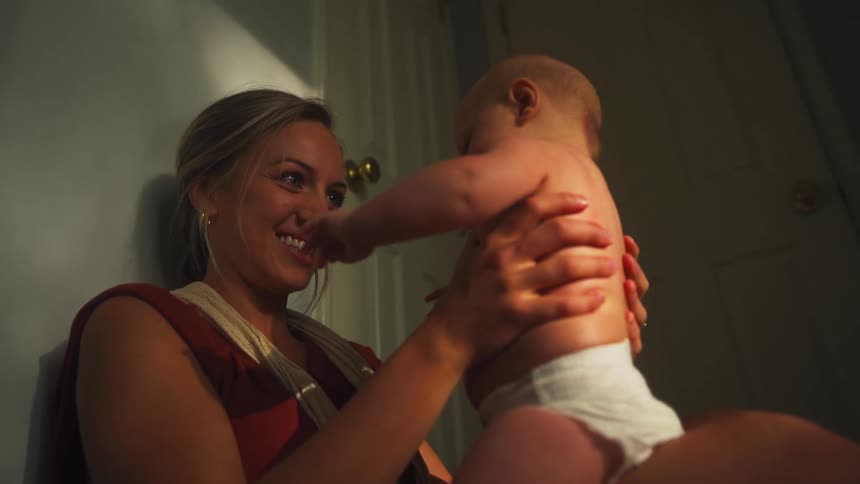A mother smiles as she holds and plays with her baby in a softly lit room at night