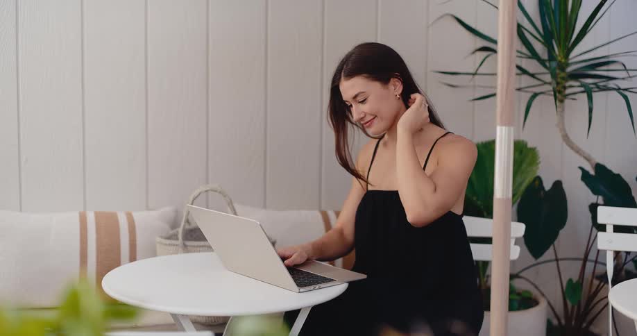 A young woman in a black dress smiles while working on her laptop at a bright, minimalist patio cafe