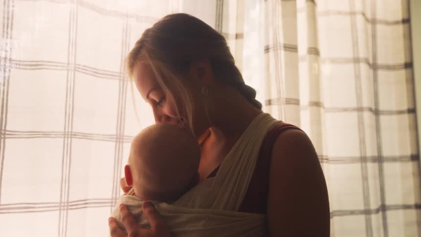 A joyful young mother holds her baby close in a sling, smiling and kissing their head in the warm, soft light of a window
