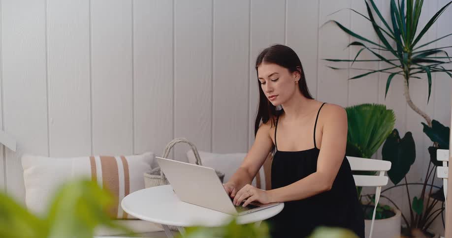 A young woman in a black dress works on her laptop while sitting on a bright, minimalist patio surrounded by plants