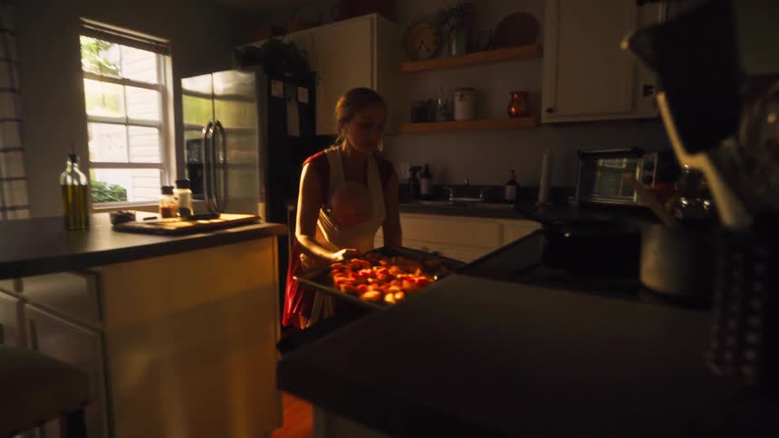 A young mother wearing her baby in a sling places a tray of freshly prepared food into the oven in a beautifully lit kitchen