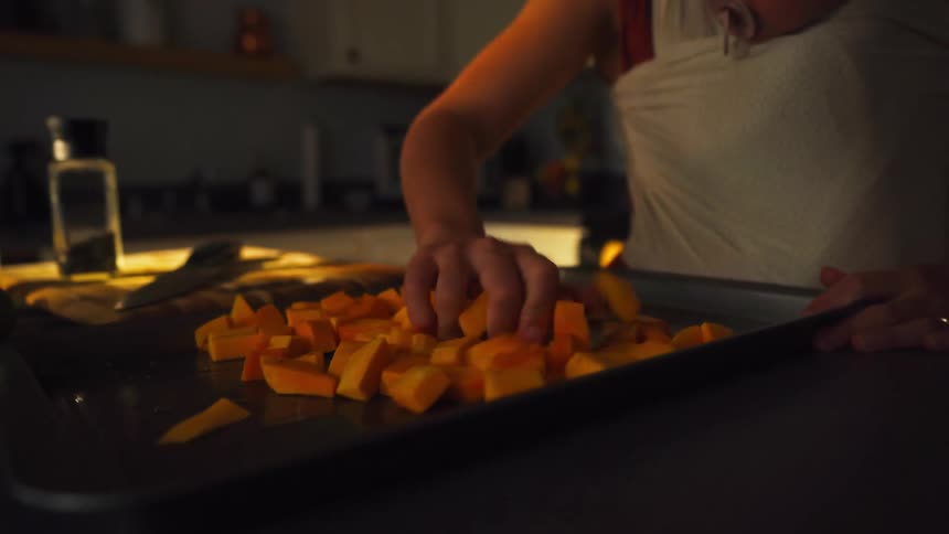 A mother prepares butternut squash for roasting in a warmly lit kitchen while her baby rests in a sling
