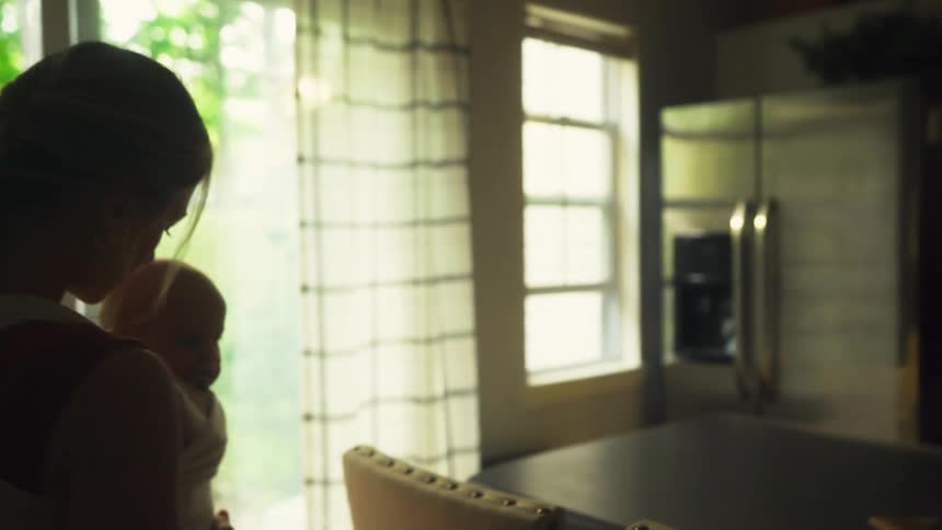 A mother with her baby in a sling turns off a warm oil lamp and walks gently through her home in the daytime