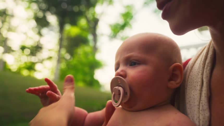 A newborn baby held in their mother's arms gently touches a leafy branch for the first time in a park