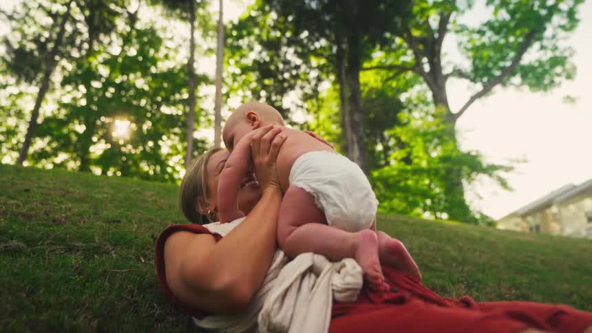 A loving mother lies on a sunny, grassy hill and playfully lifts her smiling baby boy into the air in slow motion