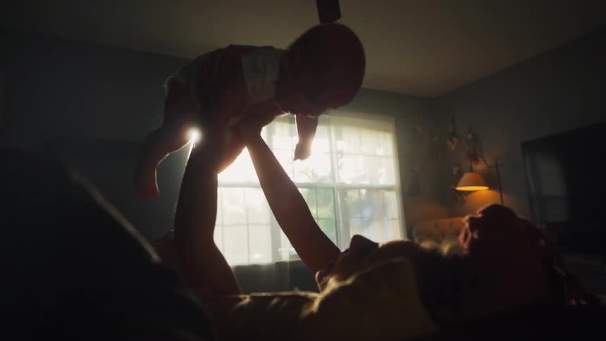 A heartwarming slow-motion shot shows a silhouetted mother playfully lifting her baby towards the bright sunlight streaming through a window