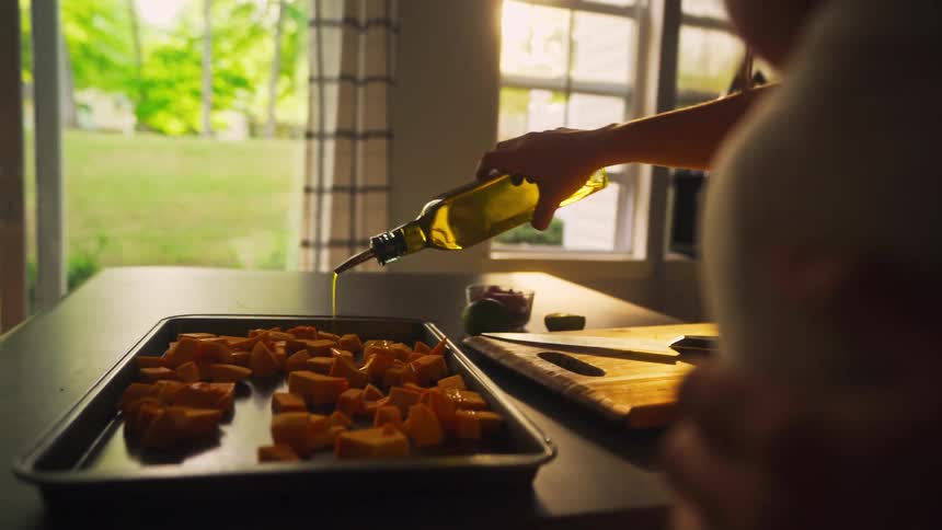 A mother, with her baby nestled in a carrier, prepares a meal in a sun-drenched kitchen, drizzling olive oil over vegetables
