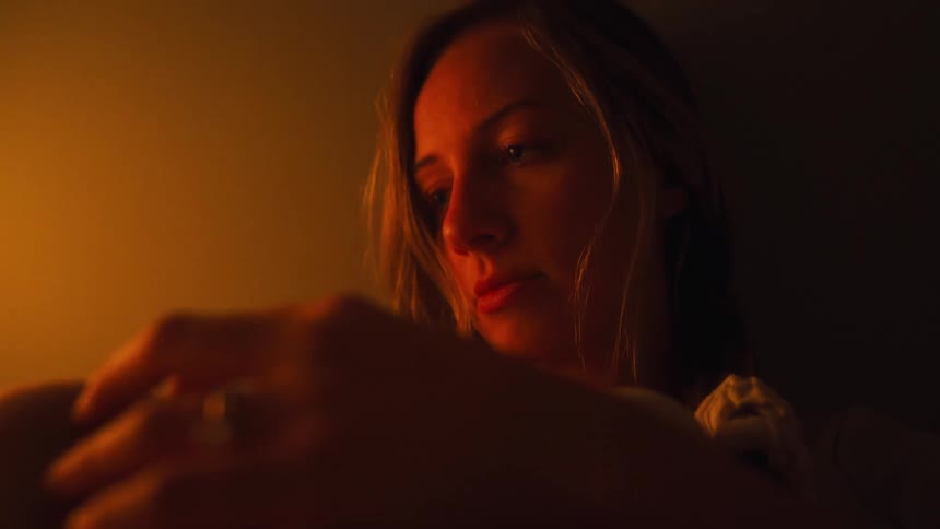 A contemplative woman gently caresses her skin while sitting alone in a dimly lit, warm room