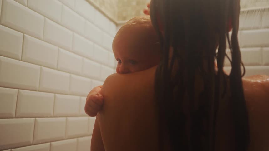 An intimate, over-the-shoulder shot of a mother holding and bathing her baby in a warmly lit shower
