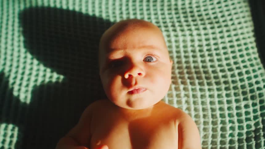 A beautiful newborn baby with blue eyes lies on a soft green blanket in the warm sunlight, looking up at the camera