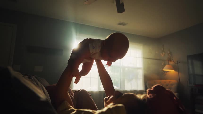 A mother joyfully lifts her baby into the air in slow motion, their silhouettes framed by the bright sunlight streaming through a window