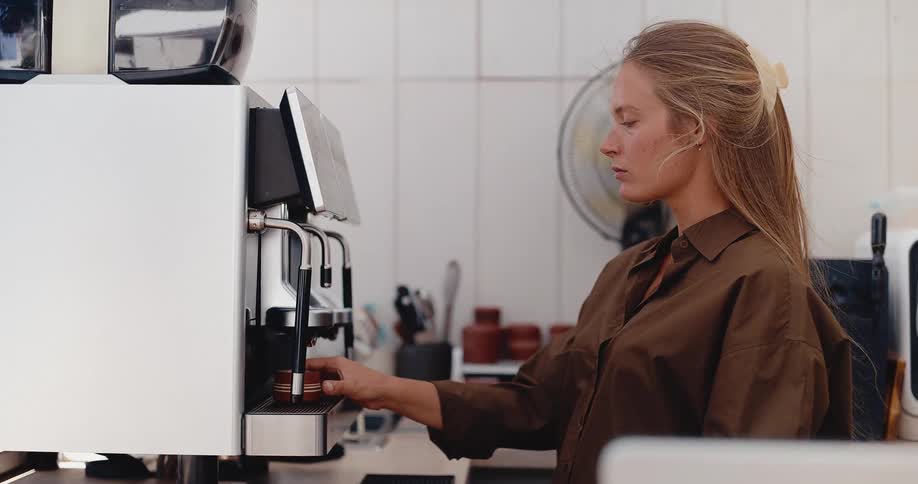 A female barista uses a modern touch-screen espresso machine to brew fresh coffee into two cups in a stylish cafe