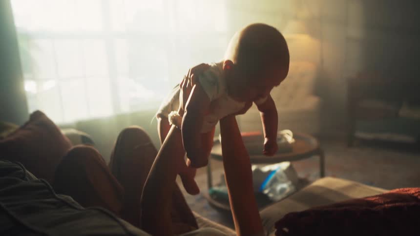 A mother gently lifts her baby up and down while relaxing on the couch in a warm, sun-drenched living room