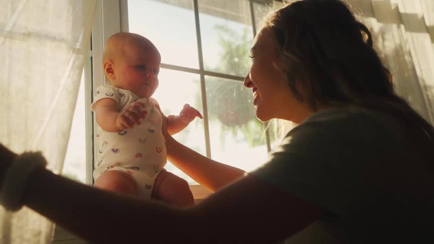 A loving mother laughs while playing peek-a-boo with her newborn baby on a windowsill, backlit by the warm afternoon sun