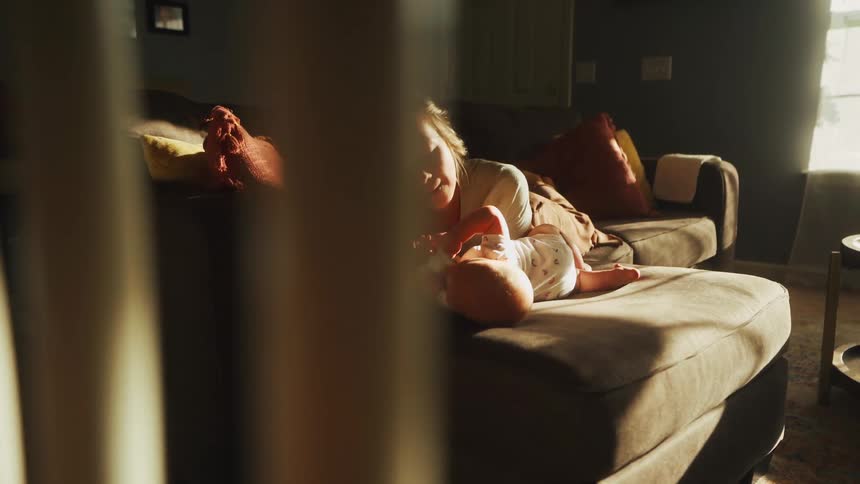 A loving mother plays with her newborn baby on the couch in a warm, sunlit living room, viewed through railings