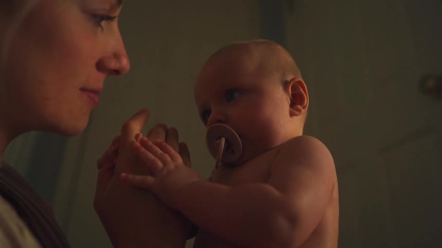 An intimate low-angle shot shows a mother lovingly holding her baby with a pacifier as they bond in a softly lit room