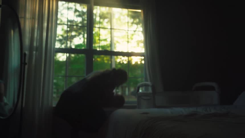 A silhouetted mother gently rocks her newborn baby to sleep in her arms as golden hour sunlight streams through the bedroom window