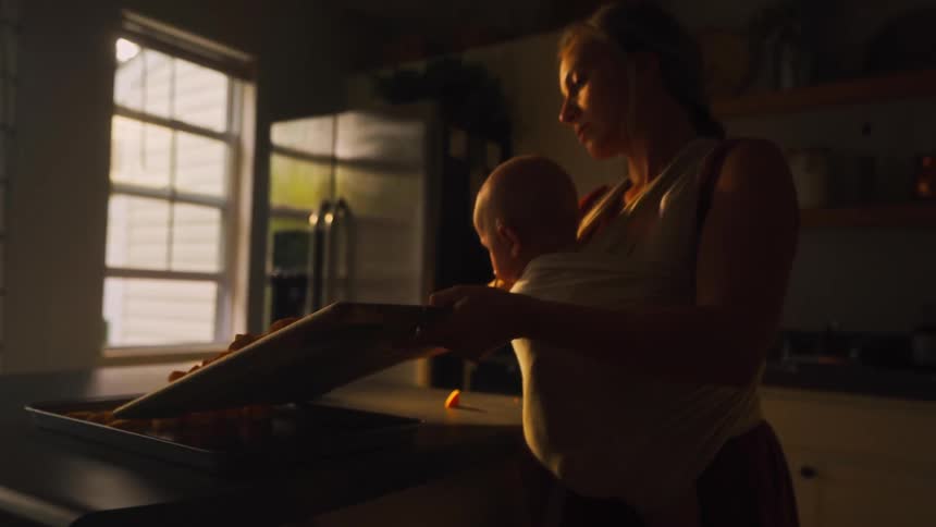 A young mother with her baby in a wrap prepares a healthy meal, sliding chopped sweet potatoes onto a baking sheet in a warm, sunlit kitchen