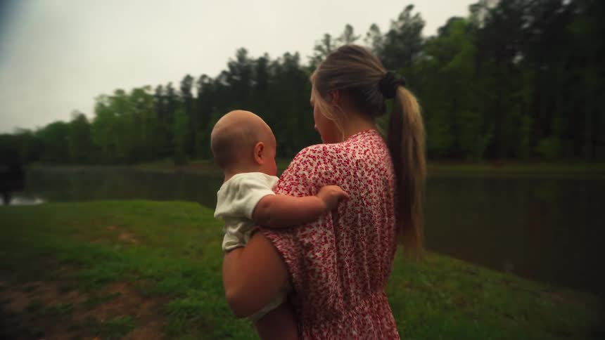 A loving mother in a floral dress gently holds her baby while walking in slow motion towards a serene lake in the woods