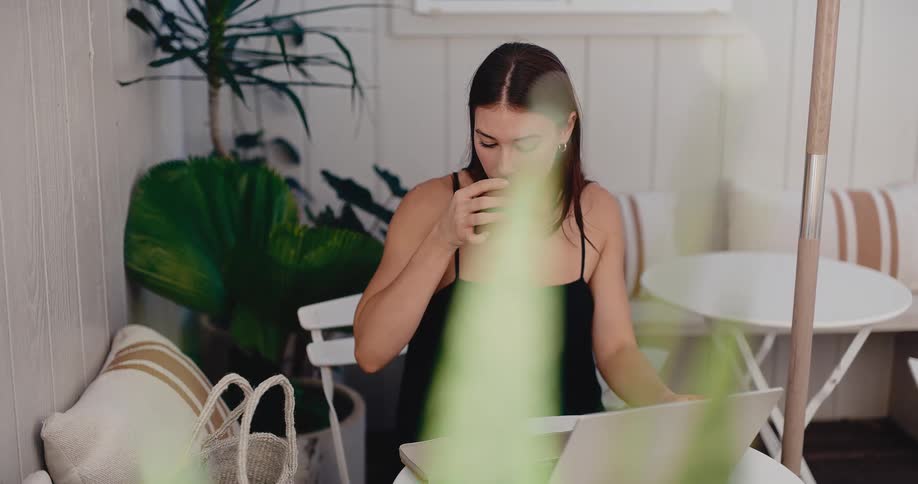 Focused woman works remotely on her laptop while enjoying a beverage on a peaceful, plant-filled patio