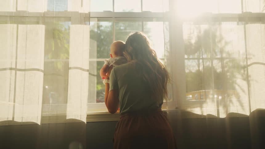 A loving mother holds her baby as they look out a sunlit window together in their home
