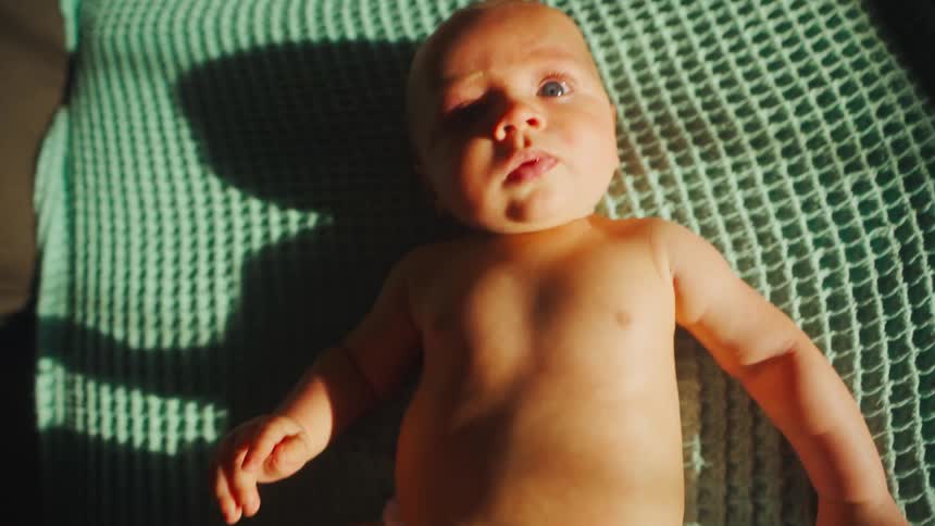 Close-up of an adorable newborn baby boy lying on his back on a green blanket, basking in the morning sunlight