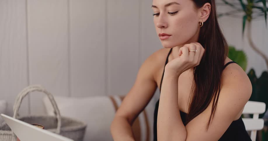 A pensive young woman works on her laptop in a bright and serene setting