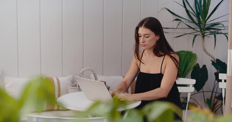 A young woman in a black dress works on her laptop, typing on a bright patio surrounded by green plants