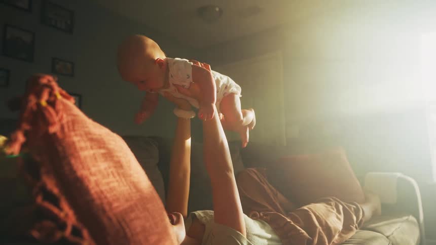 A loving mother plays with her newborn baby on the couch in slow motion, bathed in warm, golden sunlight