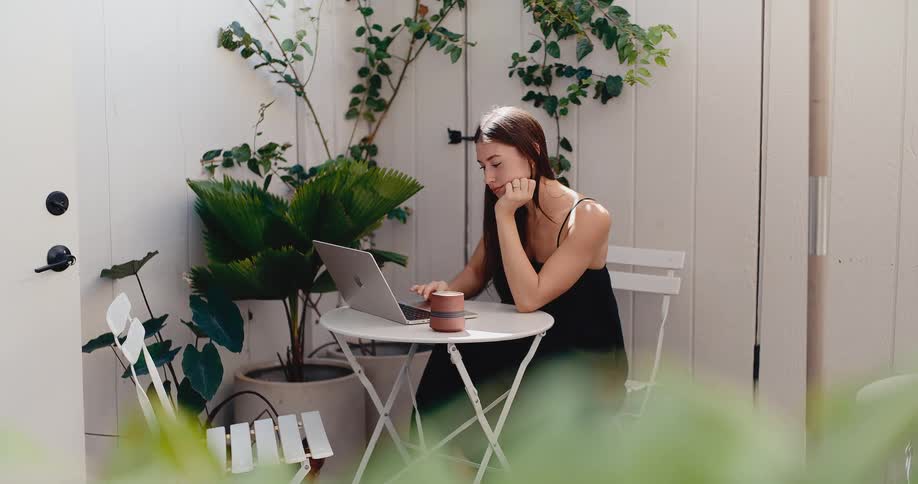 A stylish young woman works on her laptop in a tranquil, sunlit courtyard filled with lush green plants