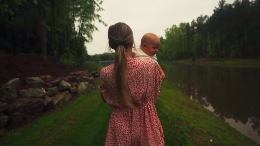 A mother in a red floral dress carries her baby while walking along the grassy shore of a serene lake in slow motion