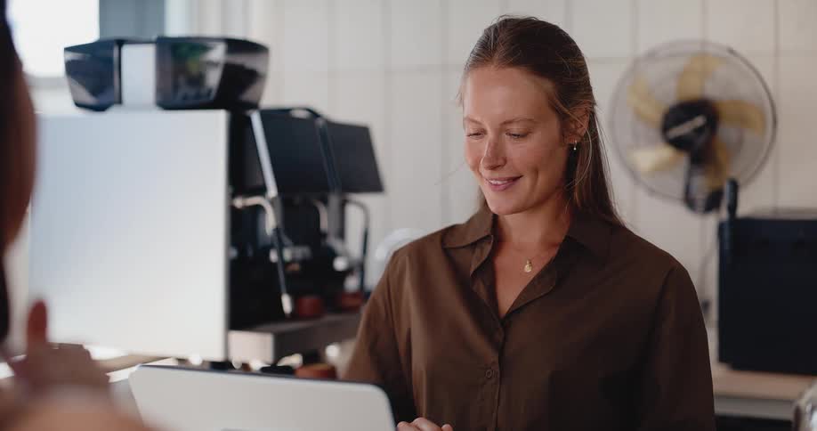 A friendly female barista with a warm smile takes an order from a customer at the counter of a bright coffee shop