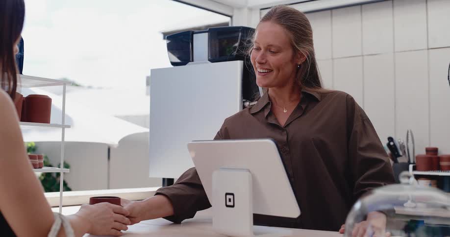 A cheerful barista smiles as she makes and hands a freshly brewed coffee to a customer in a bright, modern cafe