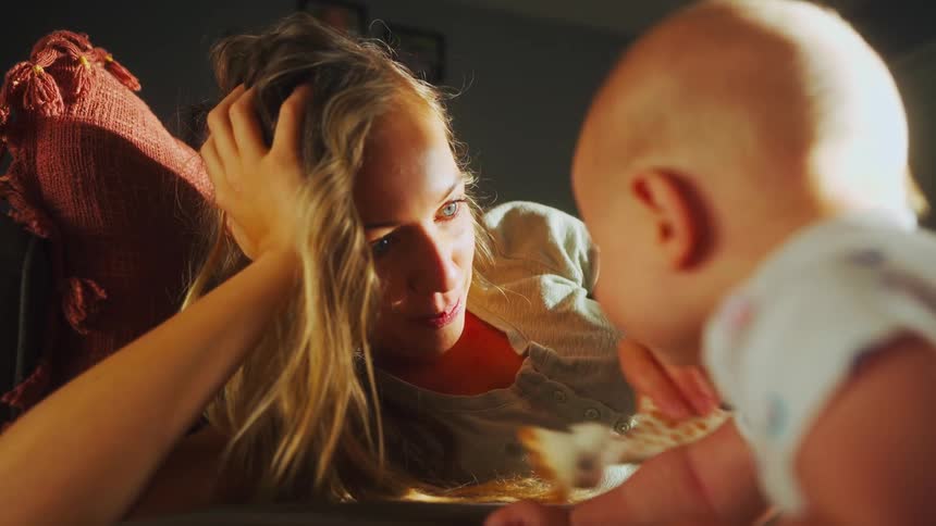 A joyful mother with long blonde hair lies on the couch and plays with her baby using a toy giraffe
