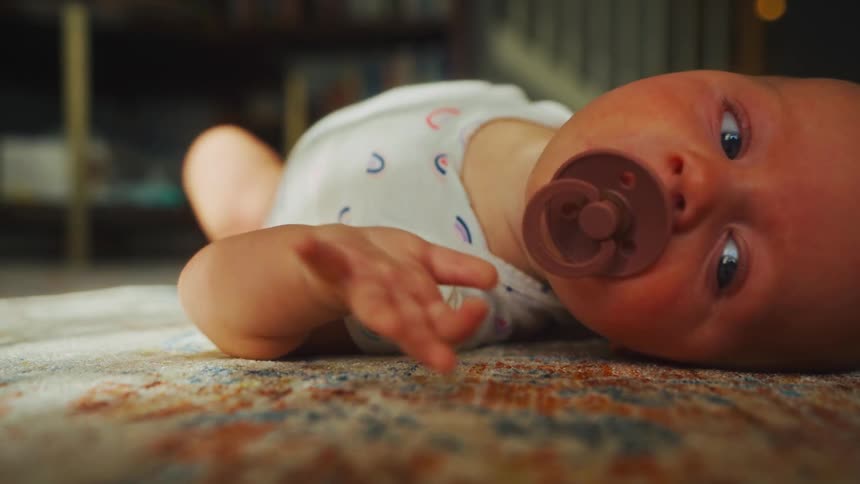 A parent's hands gently help a newborn baby roll over onto their stomach for tummy time on a patterned rug