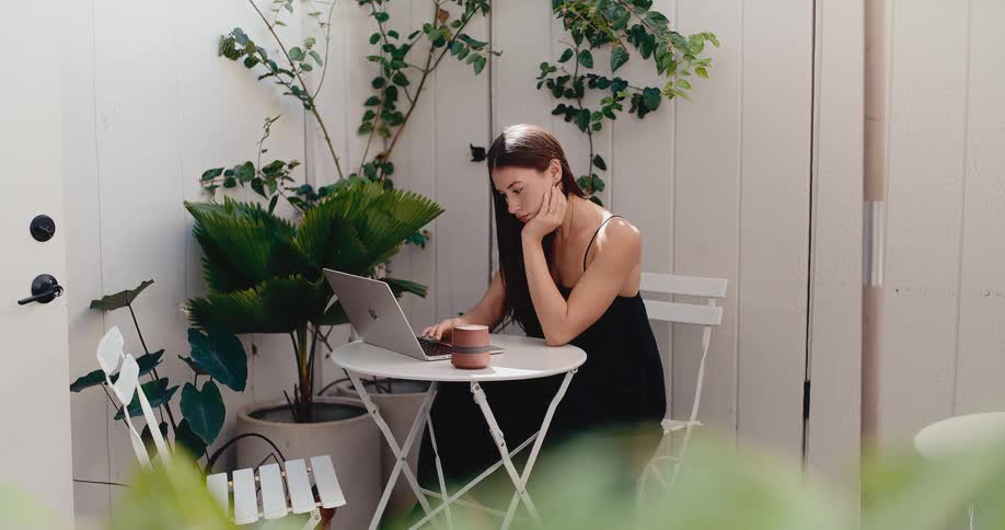 A brunette woman works remotely on her laptop, sitting at a white bistro table on a bright, sunny patio