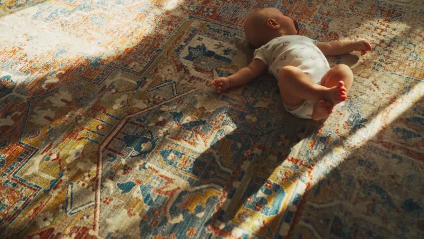 A peaceful newborn baby sleeps on a colorful patterned rug in a warm, sunlit room