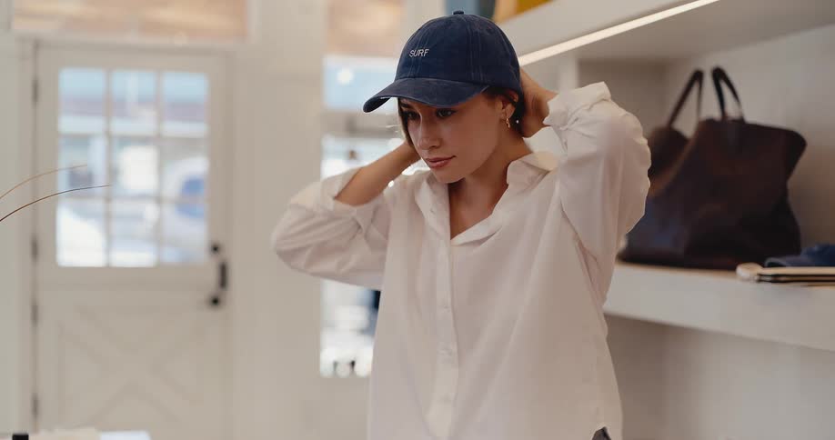 A stylish young woman slowly tries on a blue surf baseball cap while shopping in a bright, modern boutique