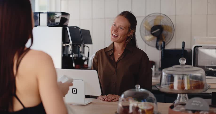 A friendly female barista smiles and assists a customer with her payment at the counter of a modern sunlit cafe